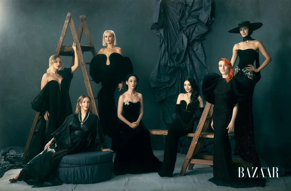 Seven women in elegant black dresses posing in a studio with ladders and draped fabric, showcasing high fashion and diverse styles in a group shot.