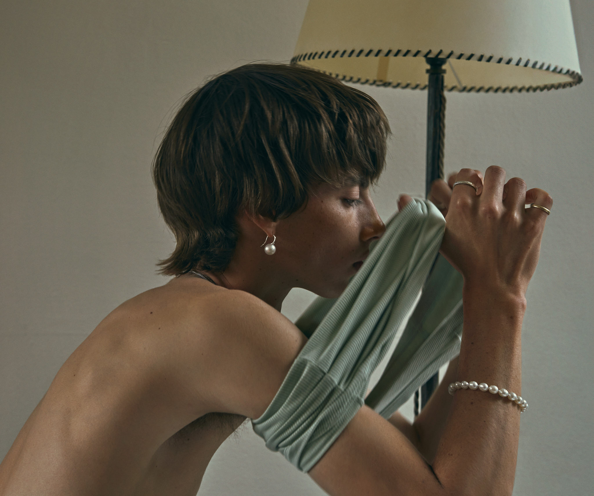 Image of a topless person with a pearl earring and bracelet sits next to a lamp wearing a light green shirt in an indoor setting.