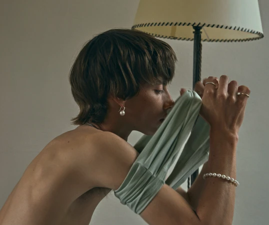 Profile of a young person pulling a sage green top over their arm, wearing pearl jewellery indoors.