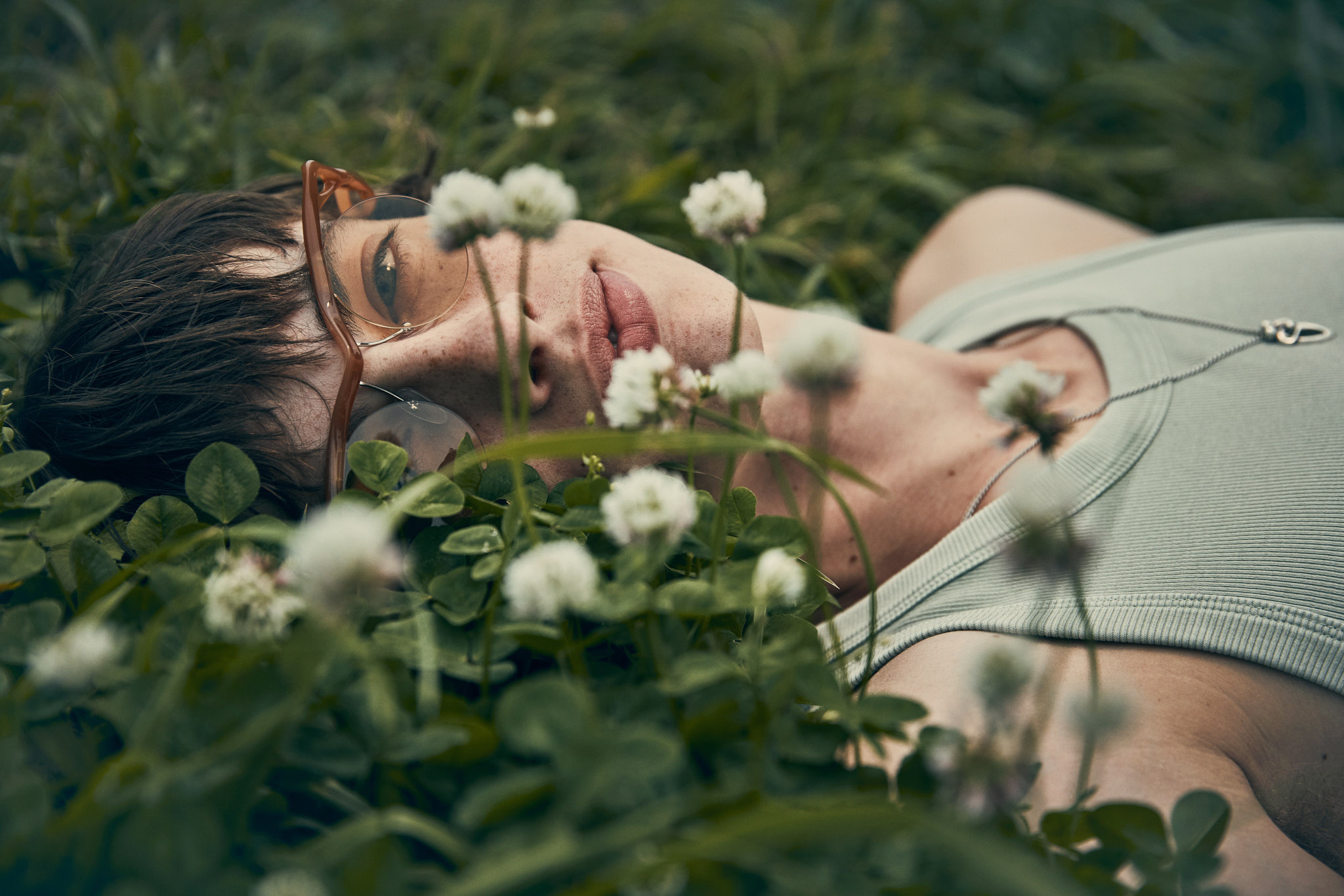 Eye-level shot of a person lying in a field of white clover flowers, wearing glasses and a light green tank top with a silver necklace.