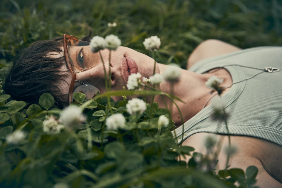 Eye-level shot of a person lying in a field of white clover flowers, wearing glasses and a light green tank top with a silver necklace.