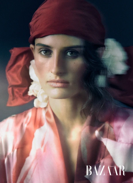 Close-up portrait of a woman wearing a red headscarf and a patterned satin robe, with 'BAZAAR' logo visible at the bottom of the frame.