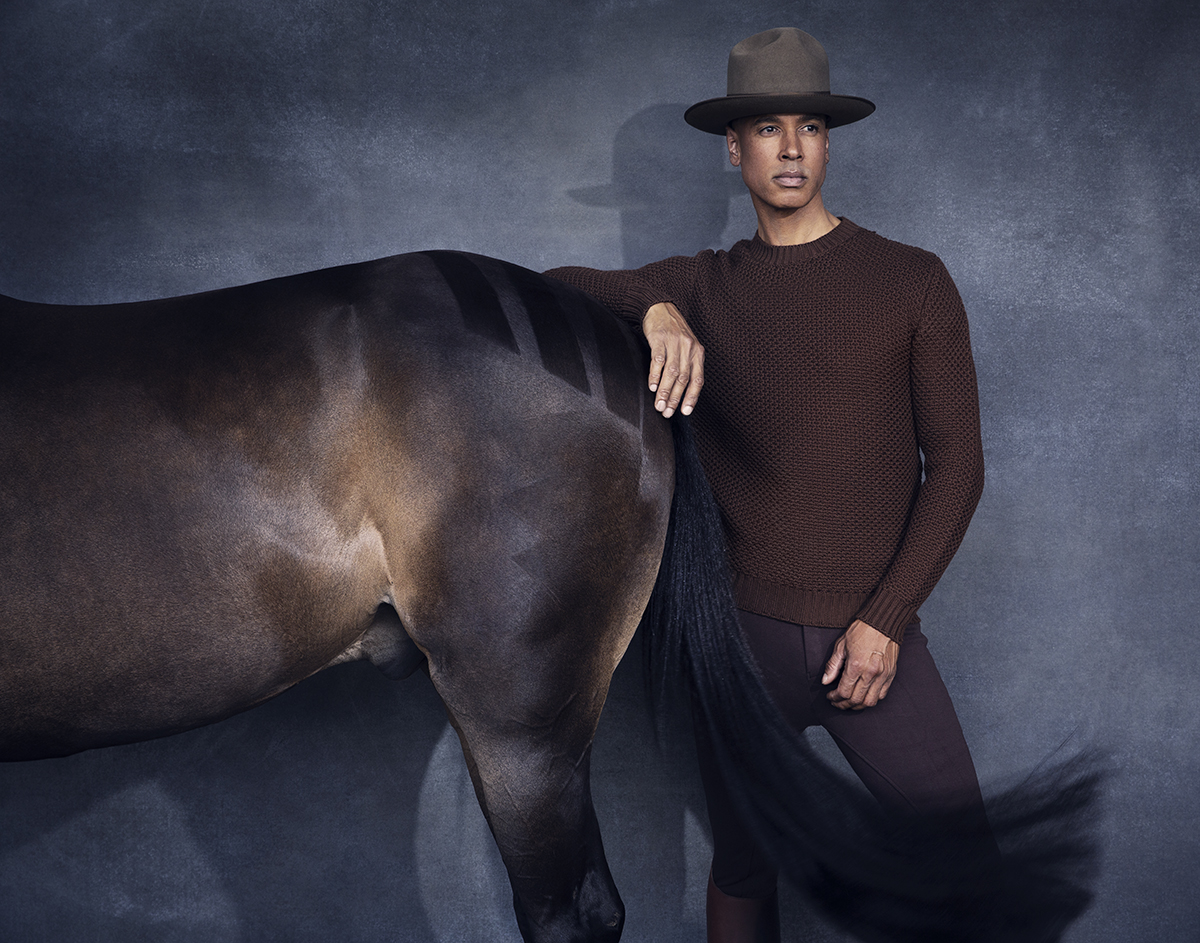Adult male in brown sweater and hat poses with a dark brown horse against a mottled grey studio backdrop; equestrian fashion shoot.