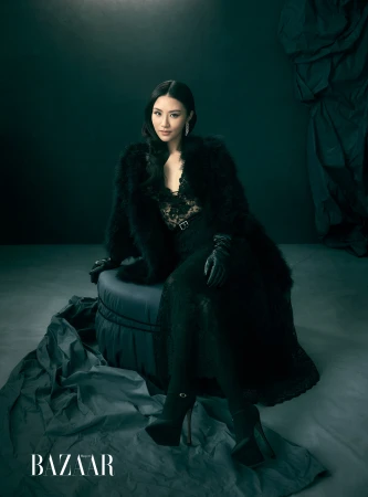 Woman in black lace dress and faux fur coat seated on a round ottoman in a dark studio