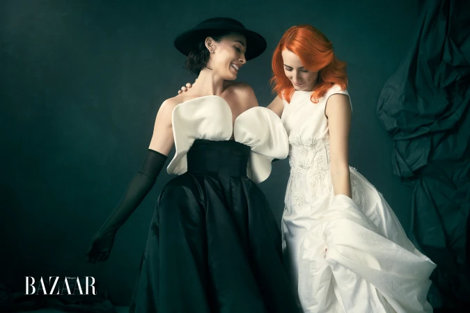 Two women laughing together, one wearing a black and white sculptural gown and hat, the other in a white dress.