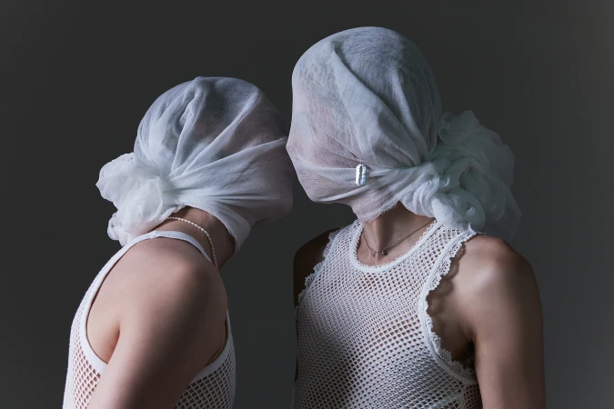 Two people wearing white mesh tank tops and draped white head coverings facing each other against a grey background.
