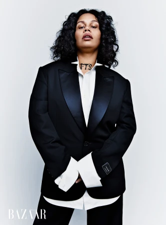 Portrait of a woman with dark curly hair wearing a black tuxedo jacket and white buttoned shirt against a white backdrop.