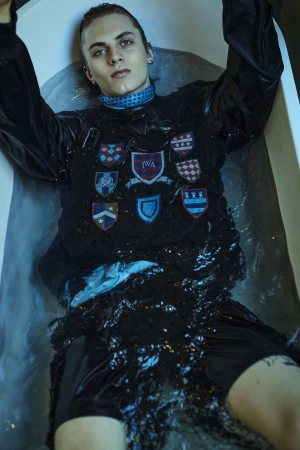 Young male model lying in a filled bathtub wearing a dark patterned top and neck wrap, shot by Michele Aboud.