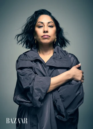 Portrait of a woman with shoulder-length wavy dark hair wearing an oversized dark grey technical jacket against a cool grey backdrop.