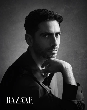Monochrome portrait of a man with dark hair and stubble resting his chin on his hand against a textured grey background.
