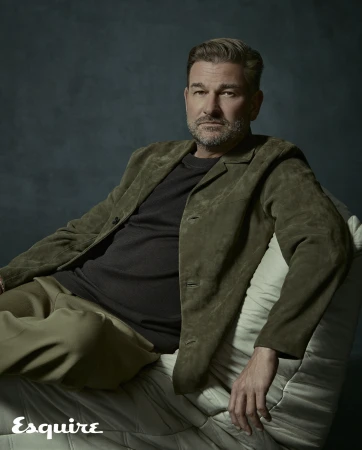 Portrait of an adult man in a green suede jacket seated against a dark textured background