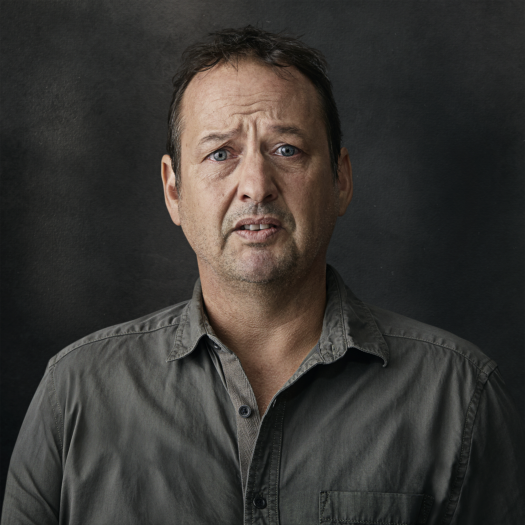 Portrait of a middle-aged man with short brown hair wearing a grey button-down shirt against a dark textured background