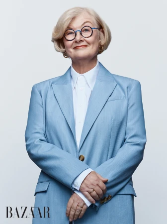 Mature blonde woman with round glasses wearing a tailored light blue blazer and white collared shirt against a white backdrop.