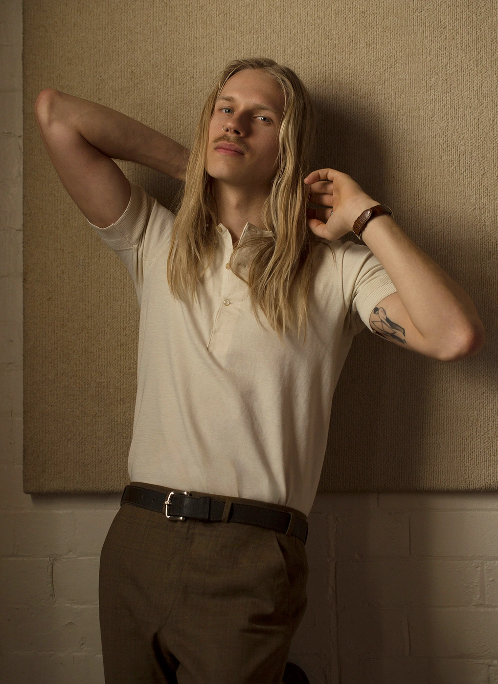 Portrait of a man with long blond hair wearing an off-white polo shirt and dark trousers against a textured panel background
