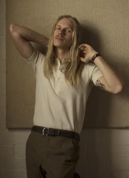 Portrait of a man with long blond hair wearing an off-white polo shirt and dark trousers against a textured panel background