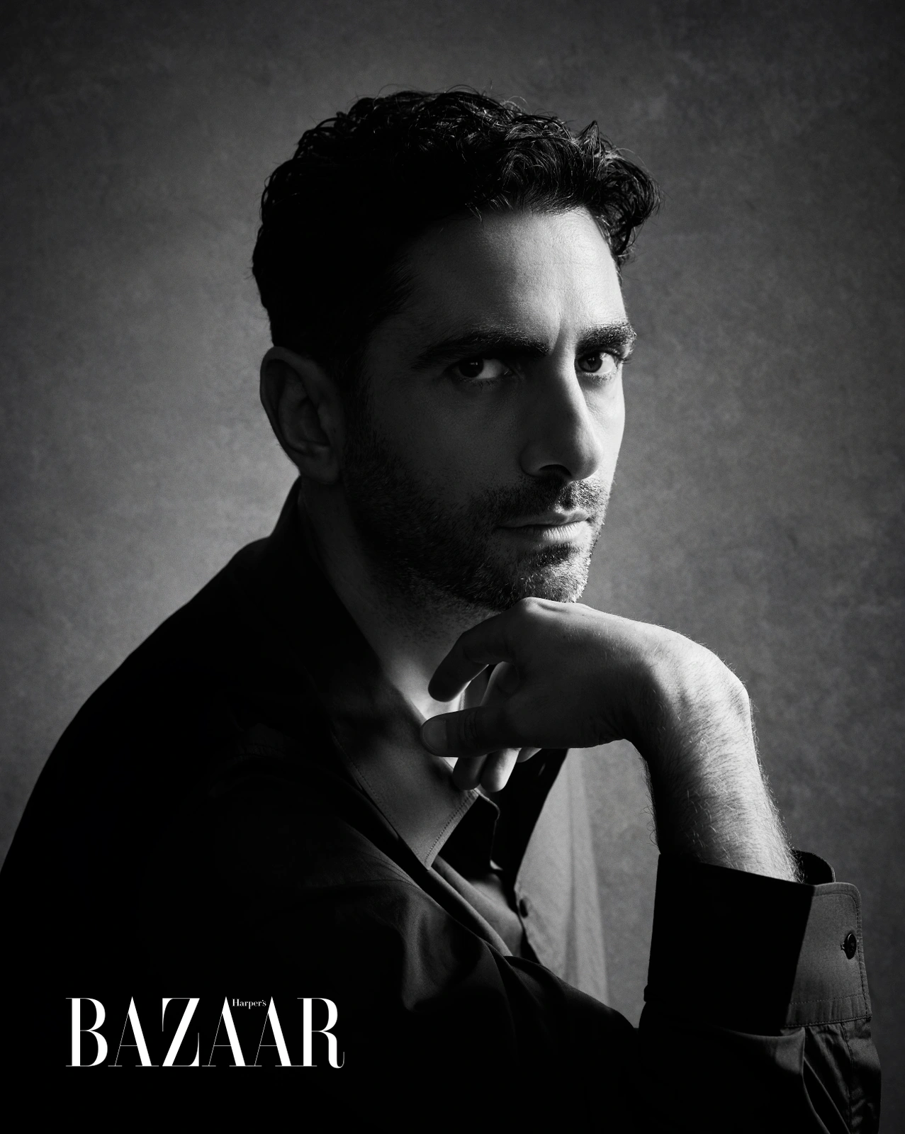 Monochrome portrait of a man with dark hair and stubble resting his chin on his hand against a textured grey background.