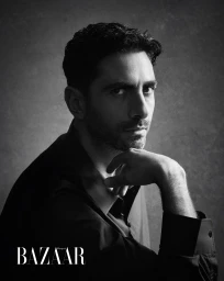 Monochrome portrait of a man with dark hair and stubble resting his chin on his hand against a textured grey background.