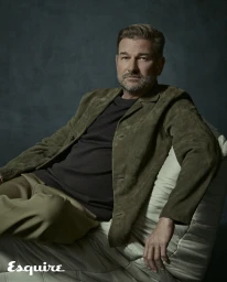 Portrait of an adult man in a green suede jacket seated against a dark textured background