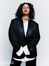 Portrait of a woman with dark curly hair wearing a black tuxedo jacket and white buttoned shirt against a white backdrop.