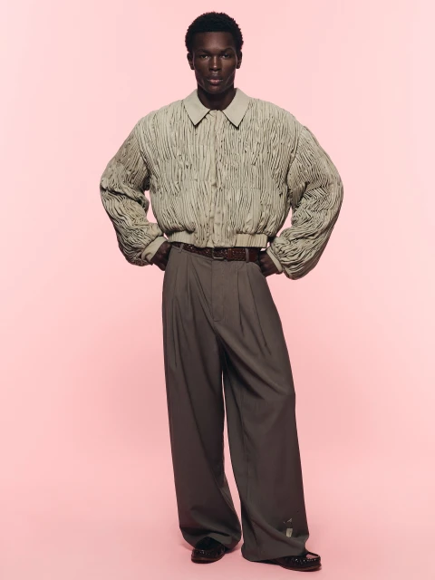 Fashionable Black male model poses confidently in a light brown bomber jacket, brown pants, and black shoes against a pink backdrop.