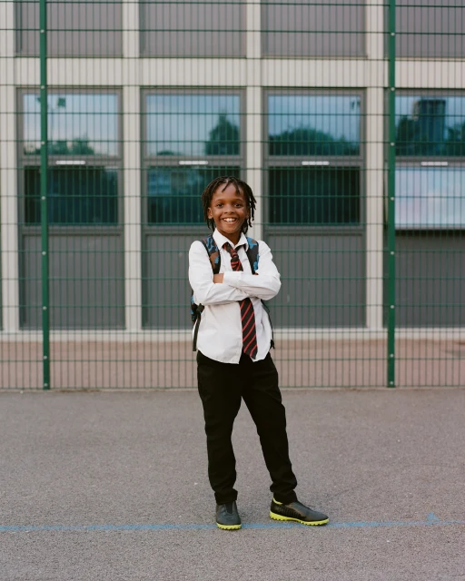 Smiling schoolboy with backpack in uniform stands outdoors with arms crossed in front of school building with bars on windows.