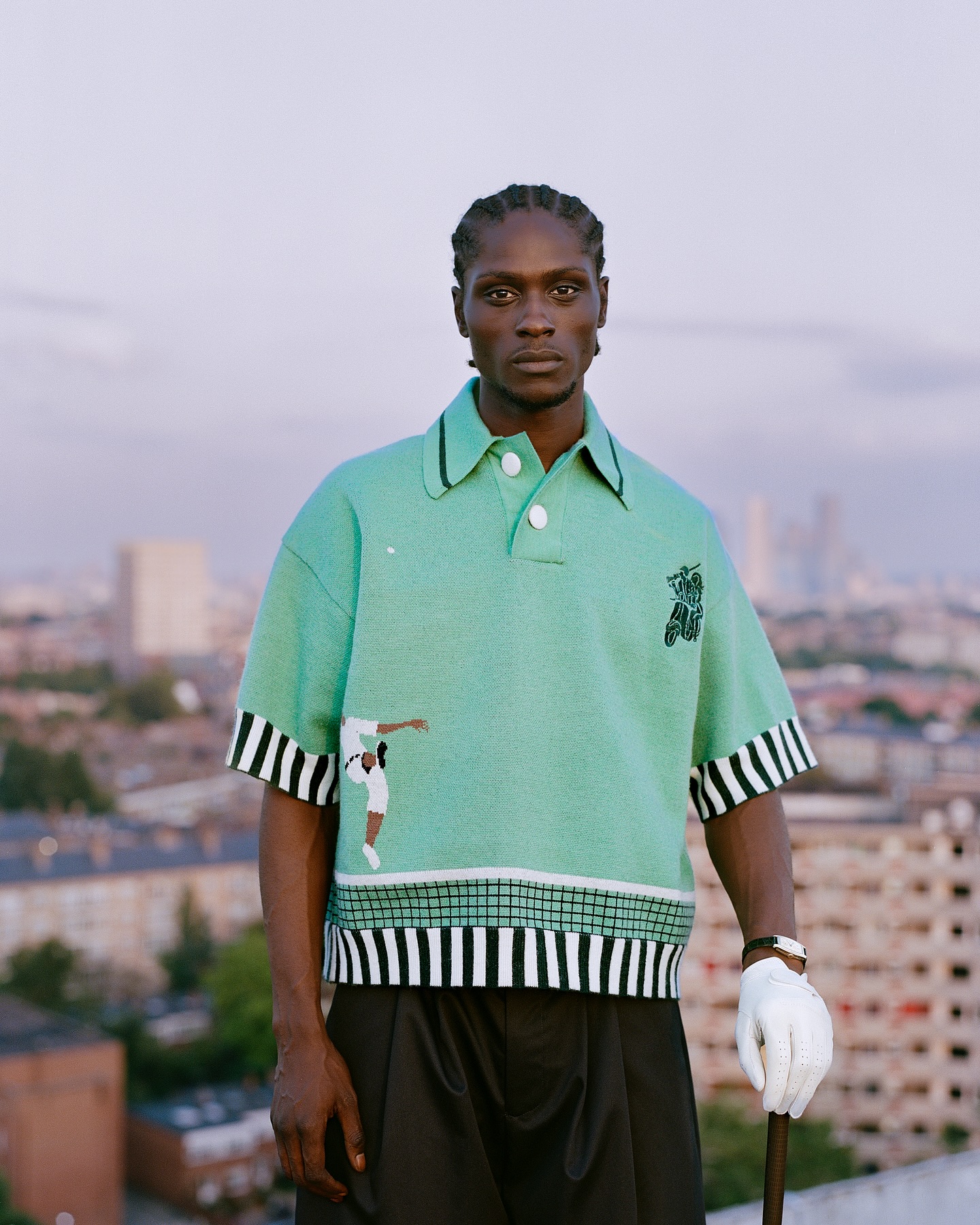 Male fashion model poses on a rooftop wearing a mint green patterned polo shirt, black pants, white glove, with a cityscape backdrop.