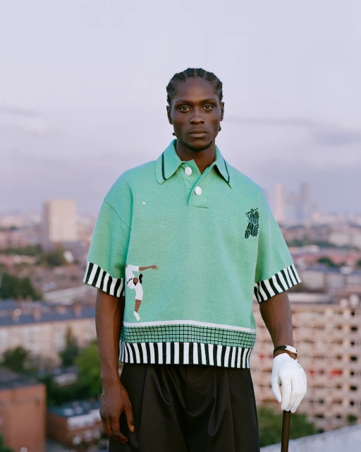 Male fashion model poses on a rooftop wearing a mint green patterned polo shirt, black pants, white glove, with a cityscape backdrop.