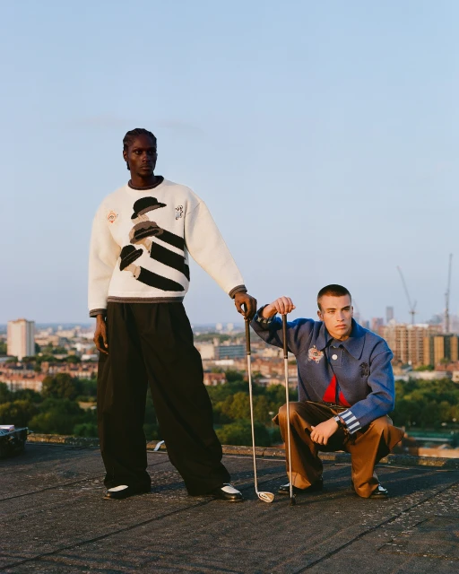 Two adult male fashion models pose on a rooftop overlooking a cityscape wearing sweaters and trousers, holding golf clubs in daylight.