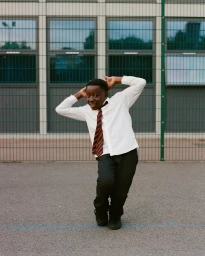 A young black boy in a white school uniform shirt and striped tie poses playfully on an asphalt school ground.