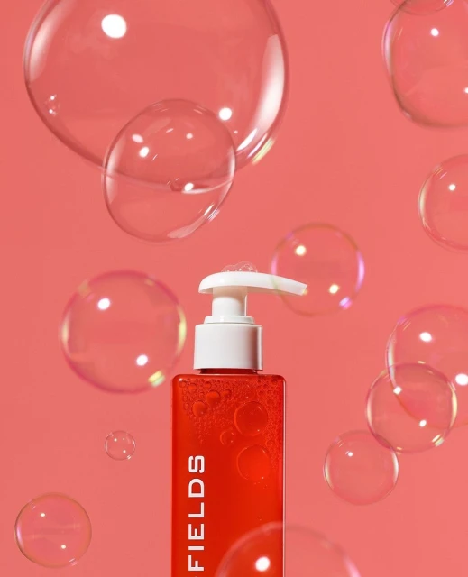 Red pump bottle of cosmetic product with white dispenser, surrounded by transparent soap bubbles on a peach-colored background.