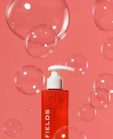 Red pump bottle of cosmetic product with white dispenser, surrounded by transparent soap bubbles on a peach-colored background.