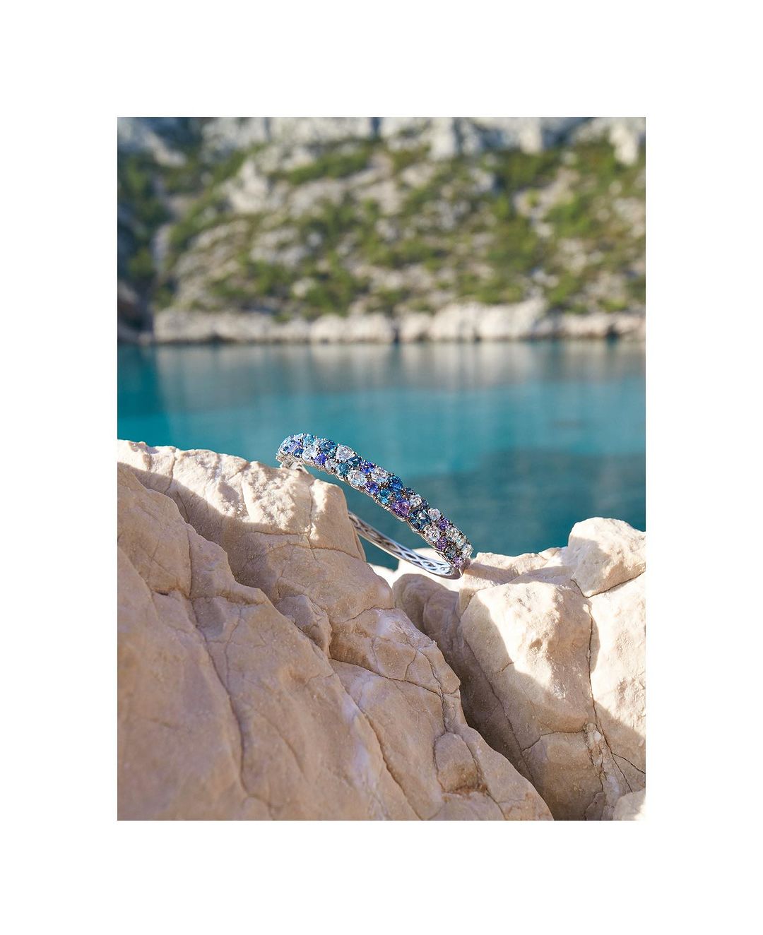 Bracelet with white and blue gems displayed on a rock with turquoise water and mountainous background, jewelry product photography.