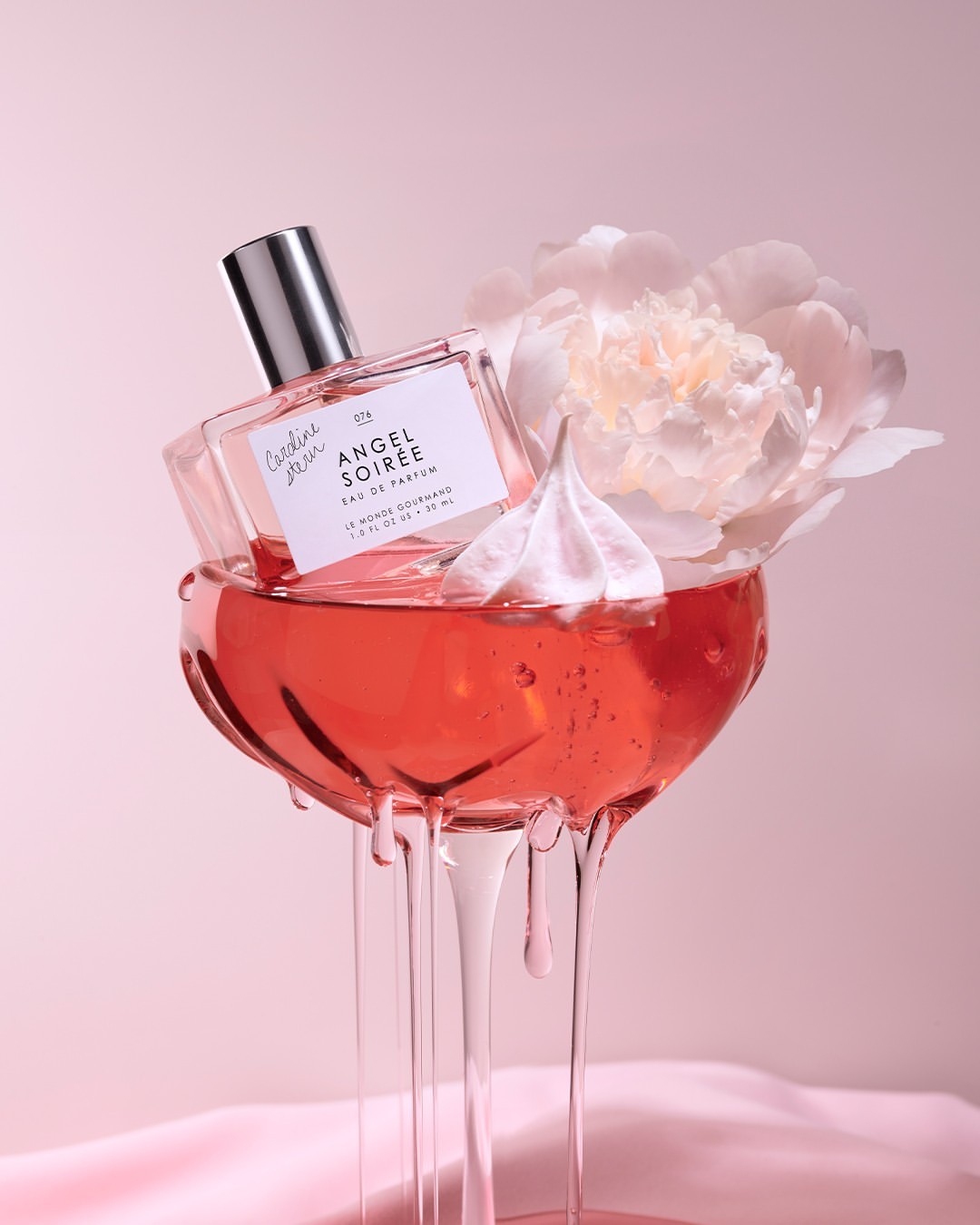 Elegant perfume bottle atop a crystal glass with pink liquid, flower, and meringue against a soft pink backdrop; beauty still life.