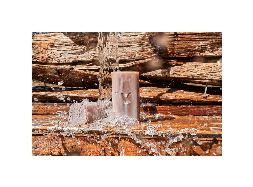 Two bars of soap sit on a wood surface as water pours over them against a background of stacked wood planks in warm lighting.