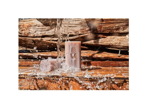Two bars of soap sit on a wood surface as water pours over them against a background of stacked wood planks in warm lighting.
