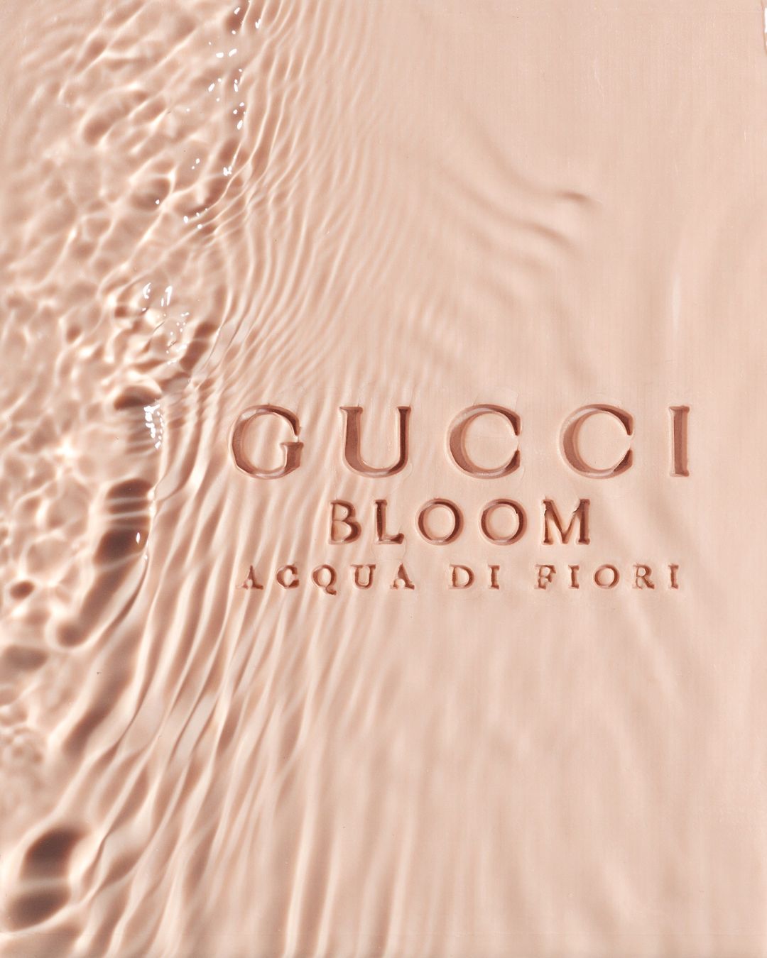Water ripples over a pink surface with the text 'GUCCI BLOOM ACQUA DI FIORI', creating a visually interesting product presentation.