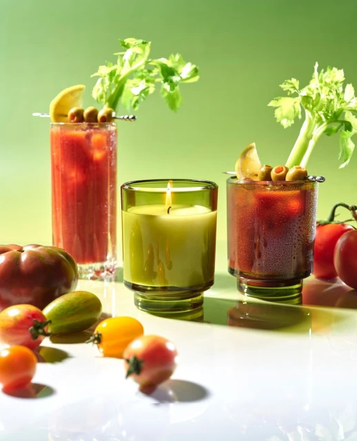Two Bloody Mary cocktails garnished with celery, olives, and lemon slices, alongside a lit candle, and small tomatoes on a white surface.