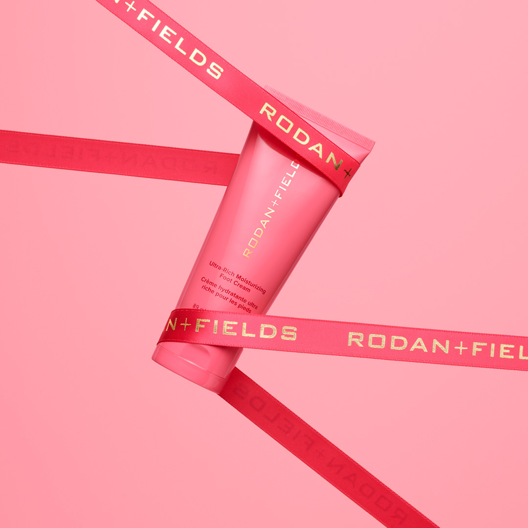 Rodan + Fields foot cream displayed with a decorative pink ribbon on a pink background. Focus on skincare product and brand.