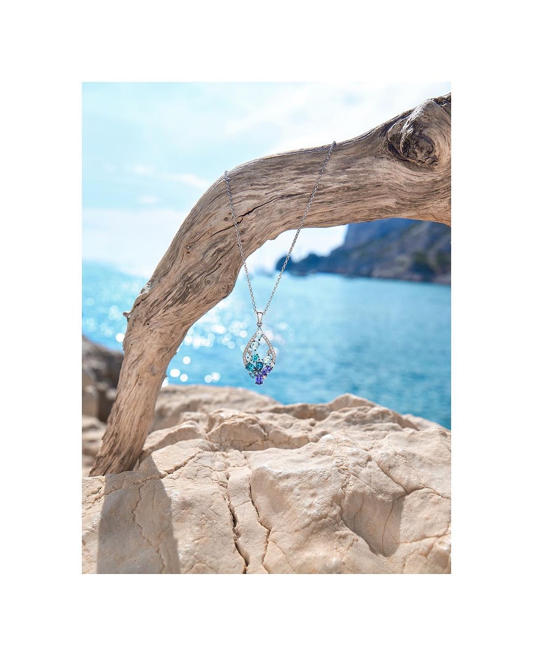 Teardrop pendant necklace with colorful gems hanging from a weathered branch, set against an ocean backdrop on a bright day.