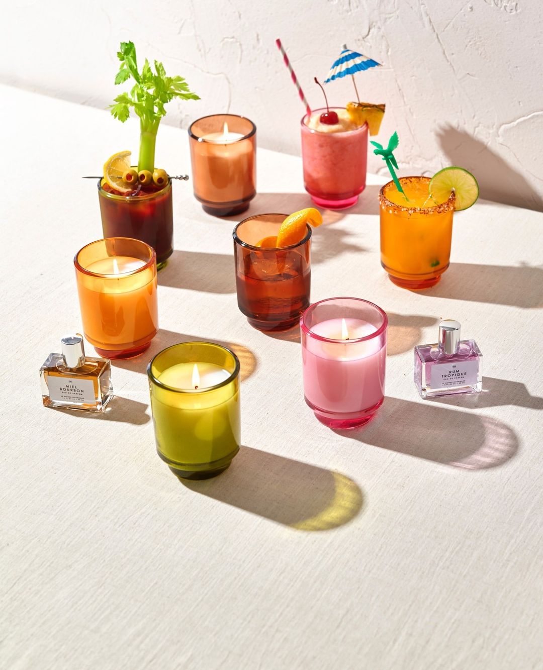 Still life of vibrant cocktail-inspired candles with decorative garnishes and perfume bottles on a white surface with diffused lighting.