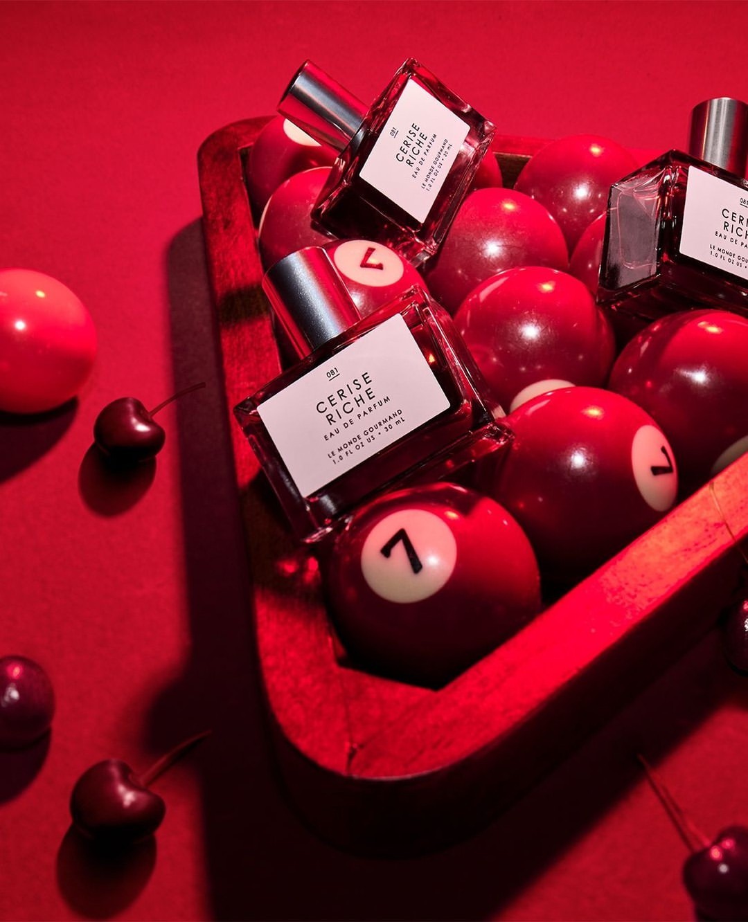 Product shot of Cerise Riche eau de parfum in a glass bottle amongst red cherries and red pool balls in a red wooden rack.