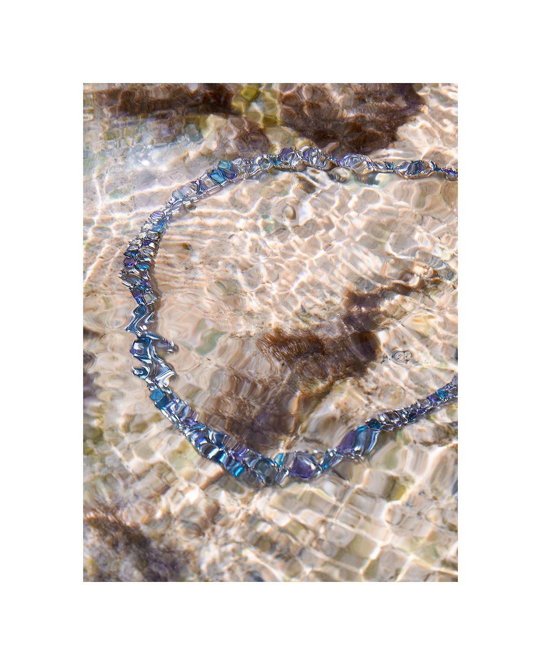 Iridescent glass beaded necklace sits partially submerged in clear, shallow water over textured brown stones in outdoor setting.