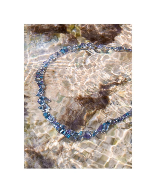 Iridescent glass beaded necklace sits partially submerged in clear, shallow water over textured brown stones in outdoor setting.