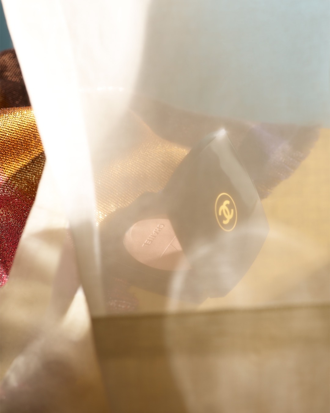 A black Chanel box with a golden logo is partially visible through a sheer white material, with colorful textiles in the background.