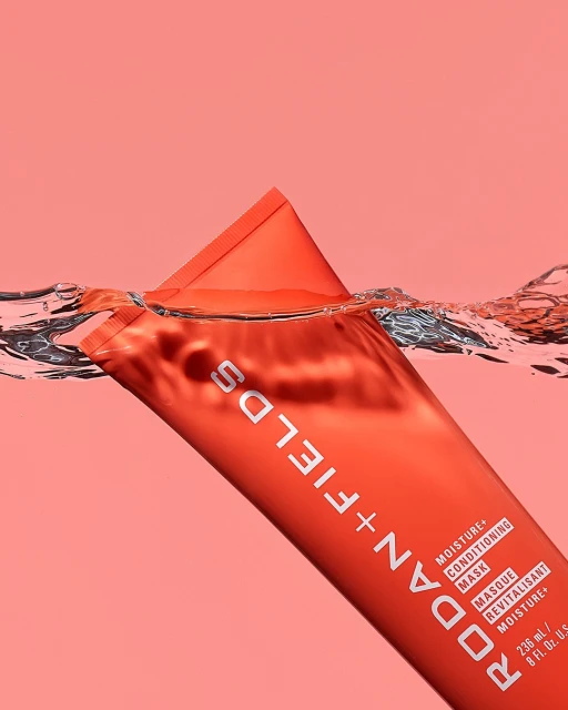 Rodan + Fields moisture mask tube at eye level emerging from water against a coral pink background in a beauty product shot.