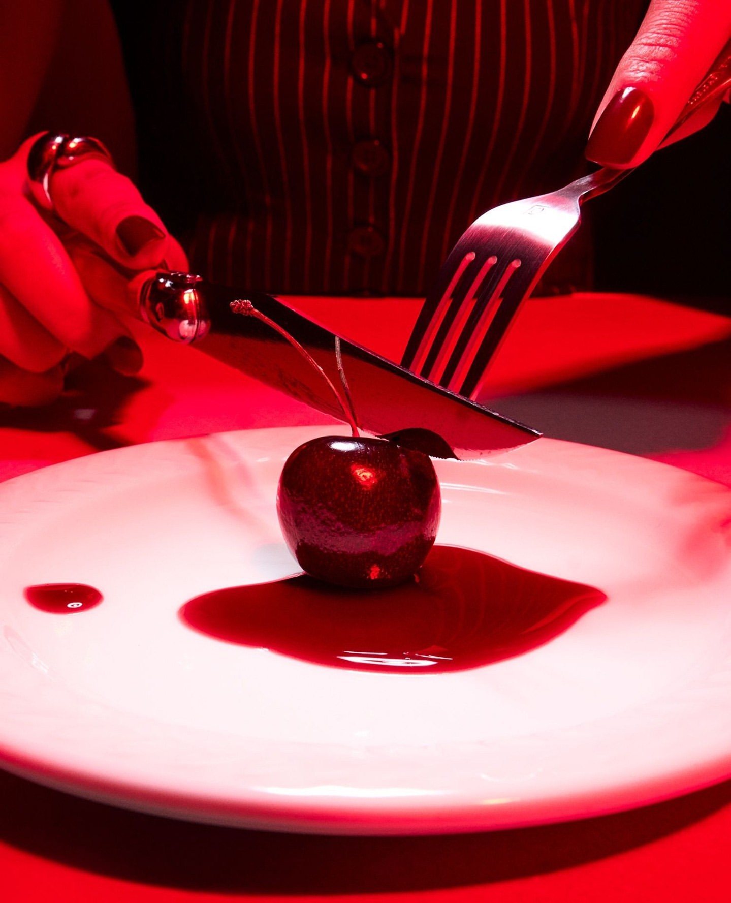 Close-up of hands cutting a glossy dark cherry on a white plate with a knife and fork under red lighting by Adam Savitch