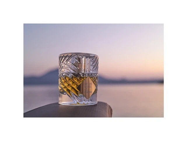 Close-up of a Kilian Paris perfume bottle, featuring clear glass and gold liquid, set against a blurred ocean and mountain backdrop at dusk.