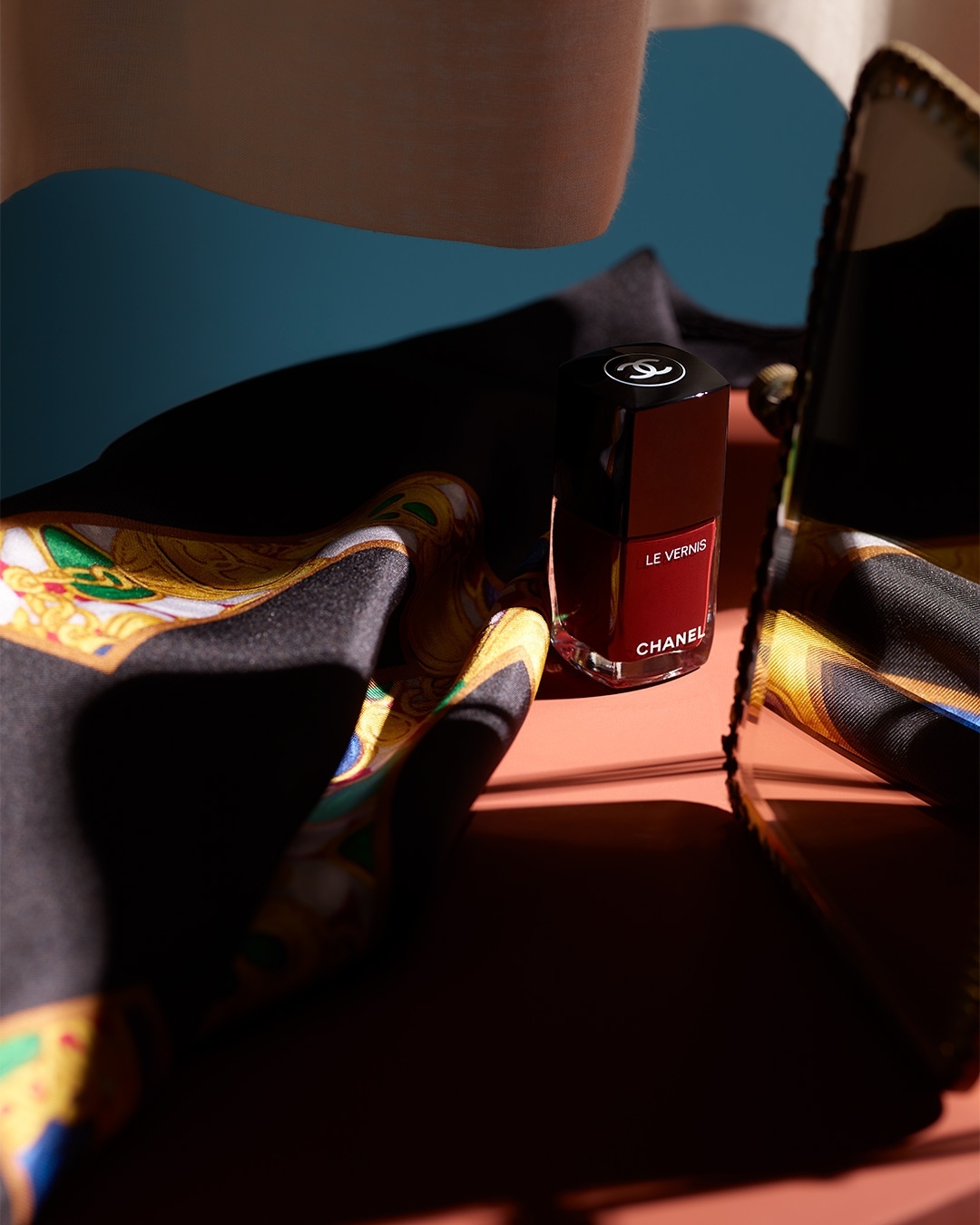 Chanel 'Le Vernis' nail polish bottle on a patterned scarf, studio setting with blue and peach backdrop, product photography.