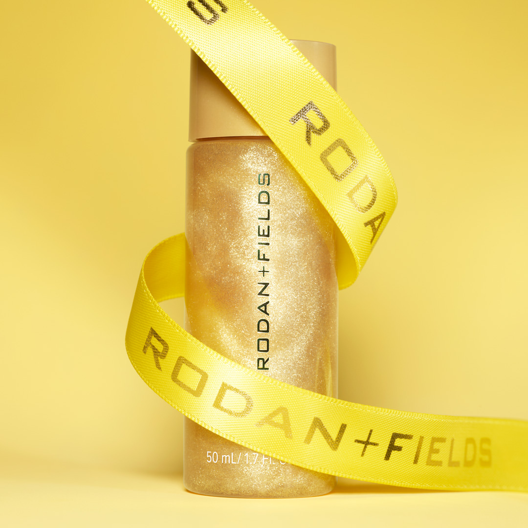 Close-up of a bottle of Rodan + Fields product wrapped with a branded ribbon against a yellow background in soft studio lighting.