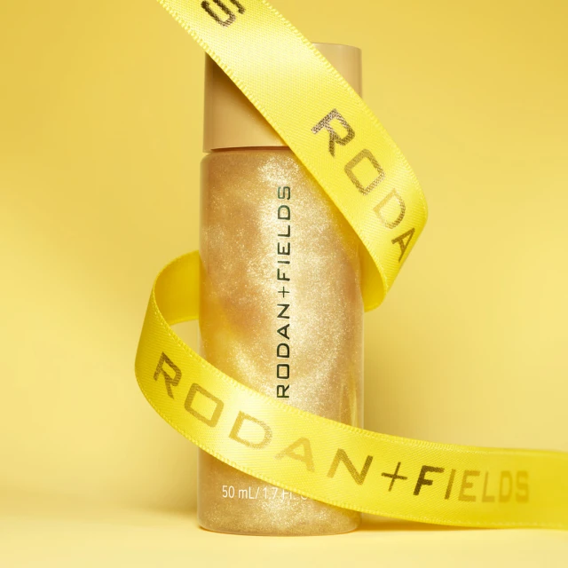 Close-up of a bottle of Rodan + Fields product wrapped with a branded ribbon against a yellow background in soft studio lighting.